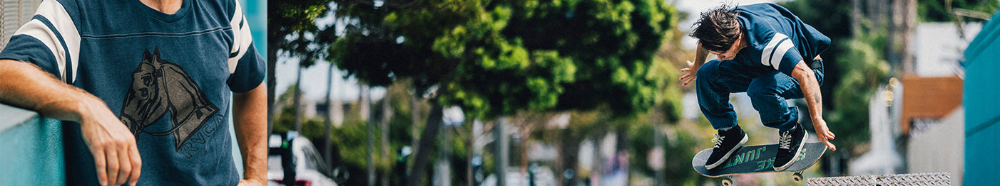 A skateboarder in blue clothes performs a trick over a bench on a city street, while another person in a blue shirt with a horse graphic stands nearby. Trees and buildings are in the background.