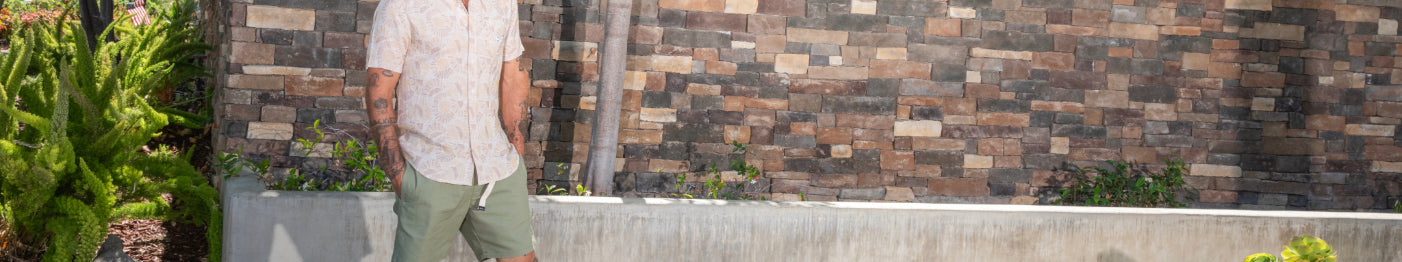 A man in a light shirt and green shorts stands outside in front of a brick wall, surrounded by greenery and plants, with sunlight casting shadows.