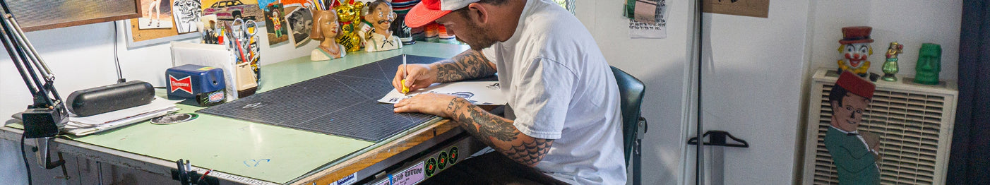 A person wearing a white T-shirt and red cap sits at a cluttered desk, drawing on paper. The workspace is filled with art supplies, figurines, and colorful decorations.