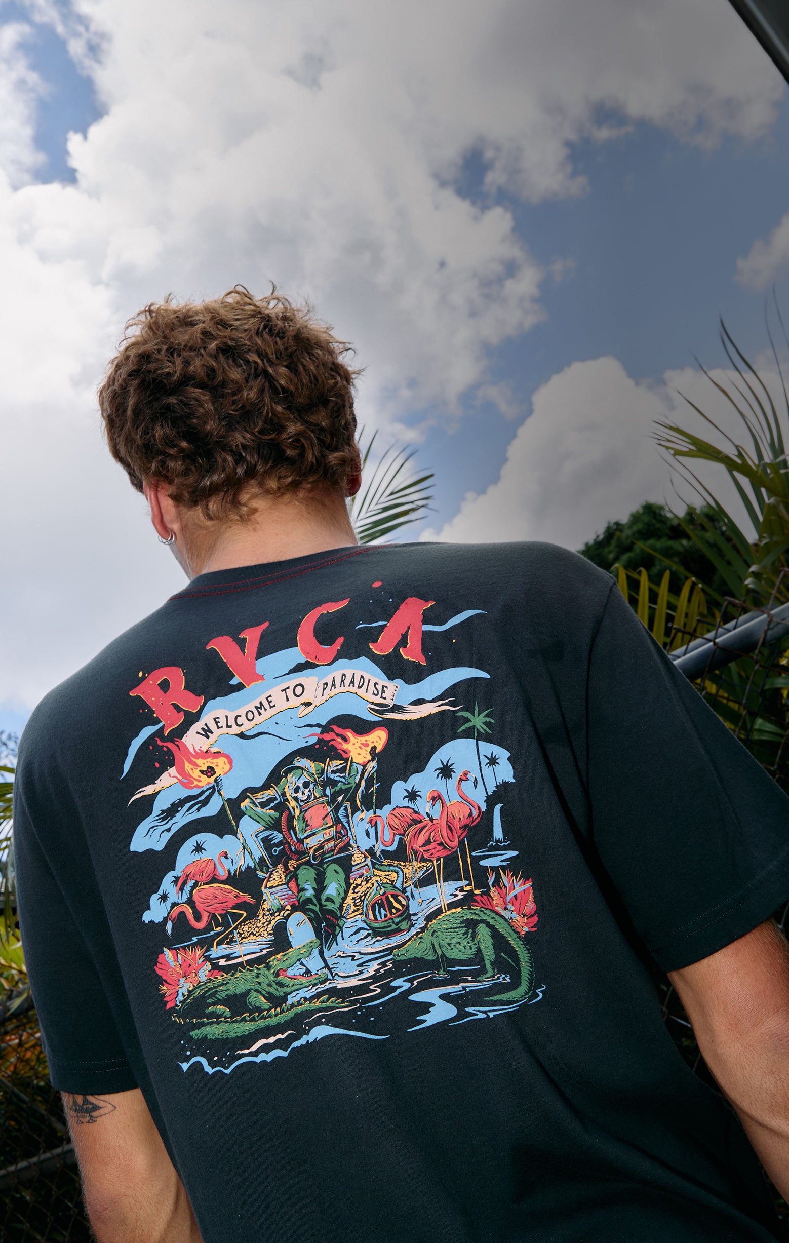 Person wearing a black t-shirt with colorful graphics and 'RVCA' branding, standing outdoors with a blue sky and clouds in the background.
