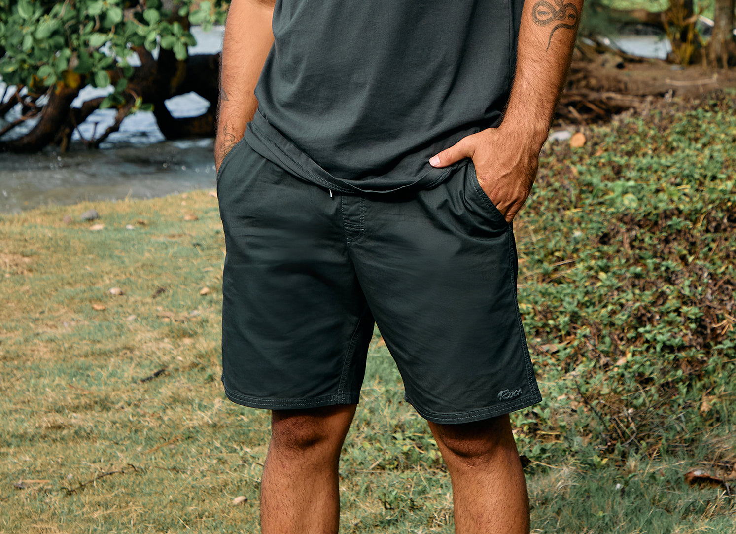 Person wearing dark green shorts and a t-shirt standing outdoors with grass and trees in the background.