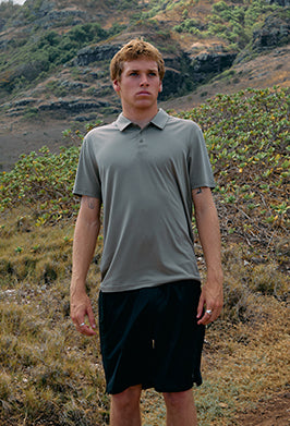 Man wearing a gray polo shirt and black shorts standing in a natural landscape.