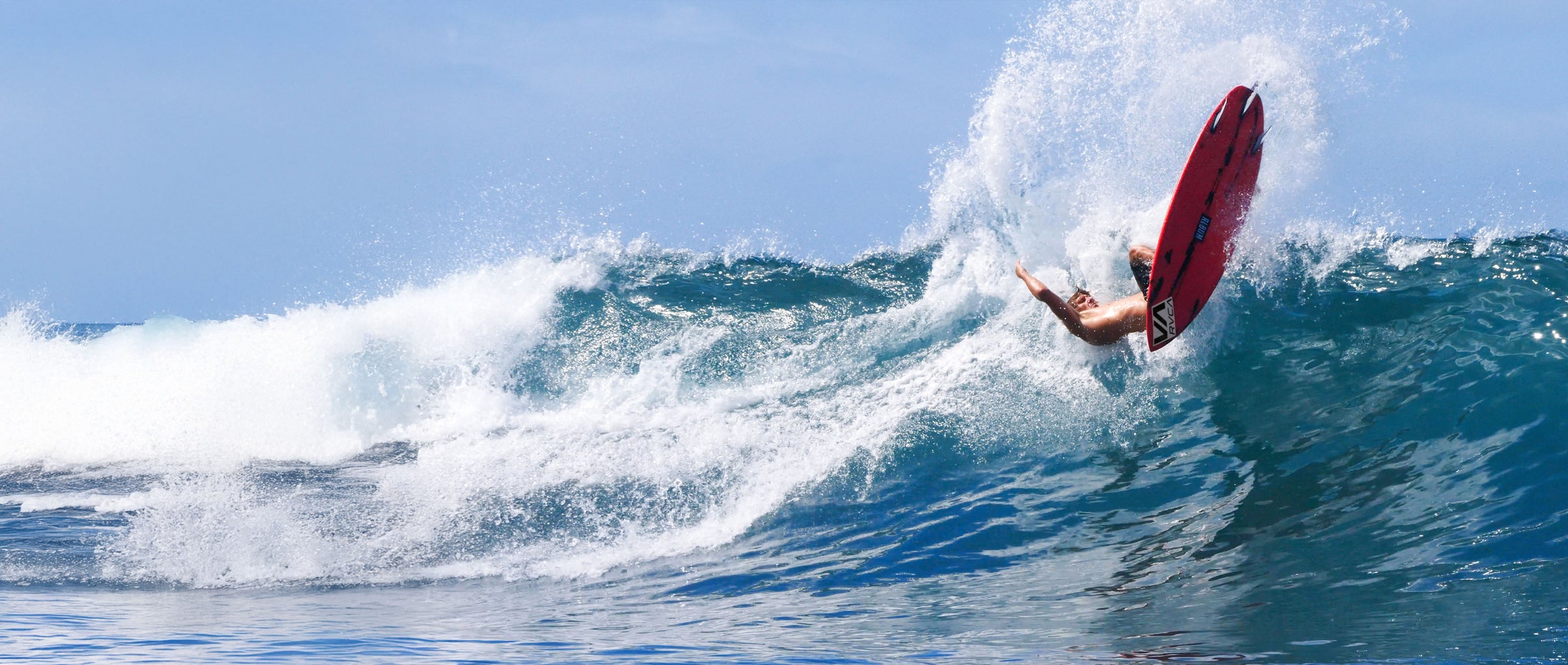 Person on a red surfboard riding a wave in the ocean with clear blue sky.