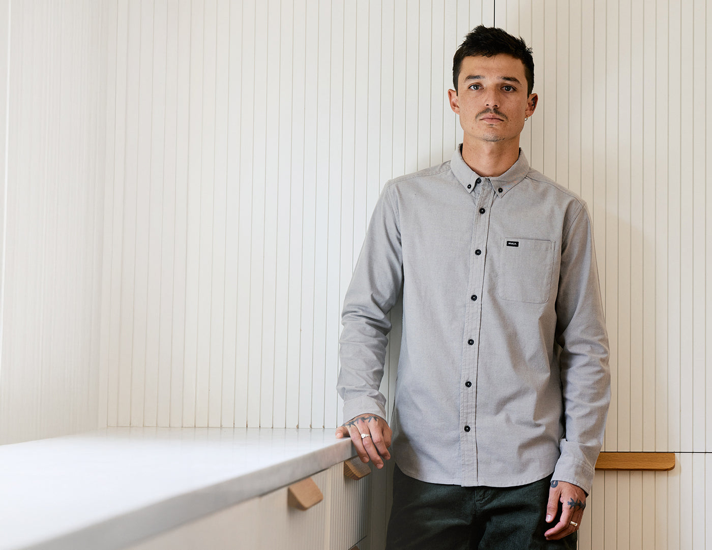Man wearing a gray button-up shirt standing against a white paneled wall.