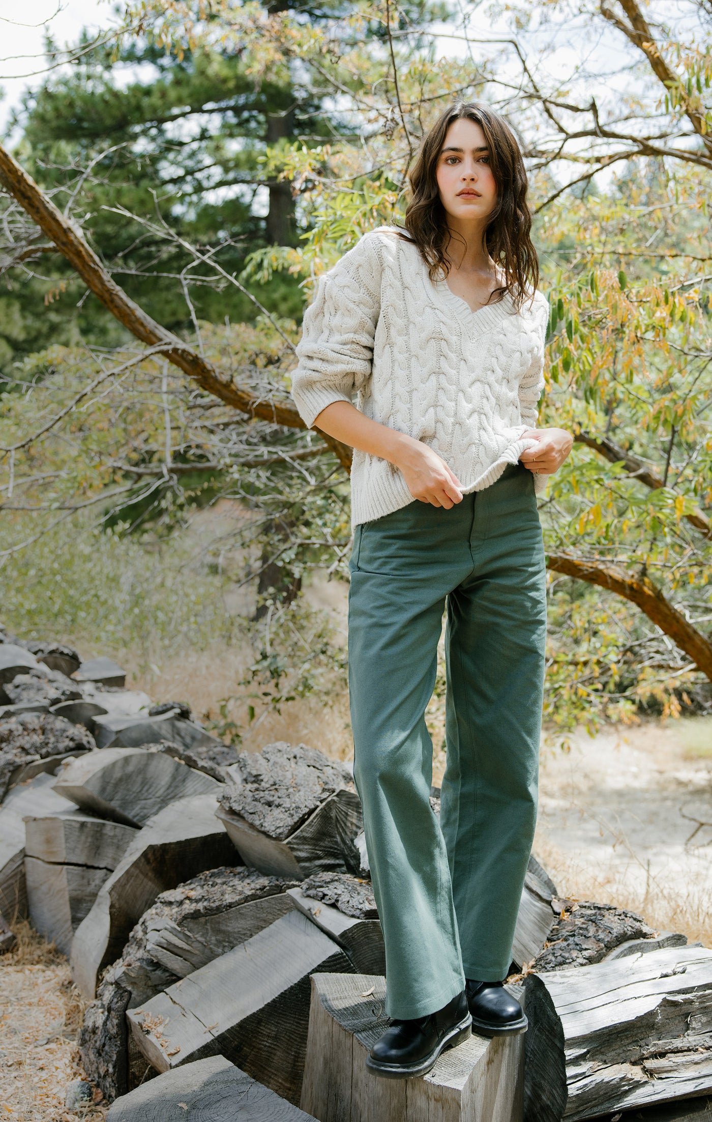 Woman standing outdoors in a natural setting wearing a light sweater and green pants.