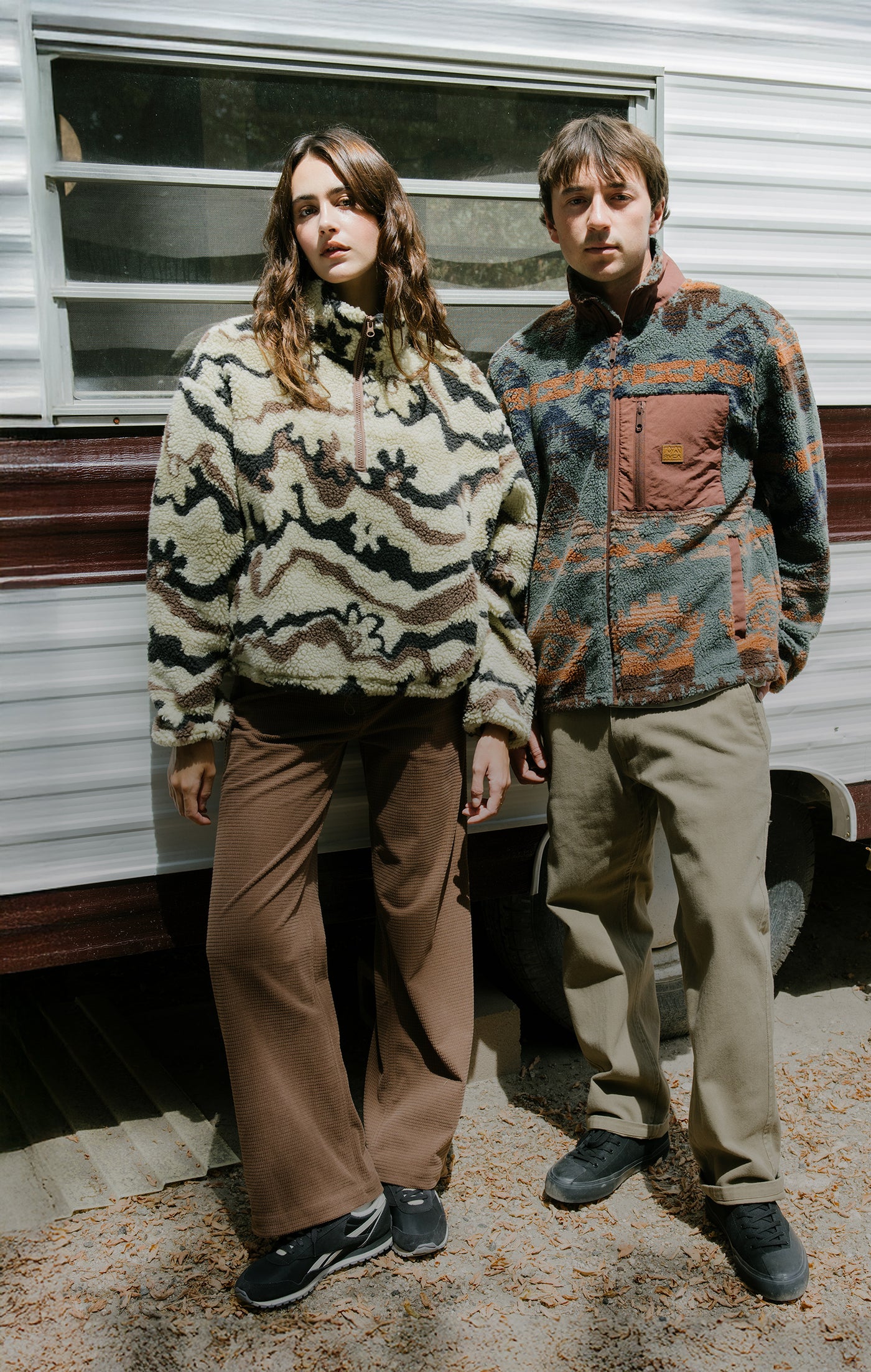 Two people standing in front of a trailer wearing patterned sweaters and pants.