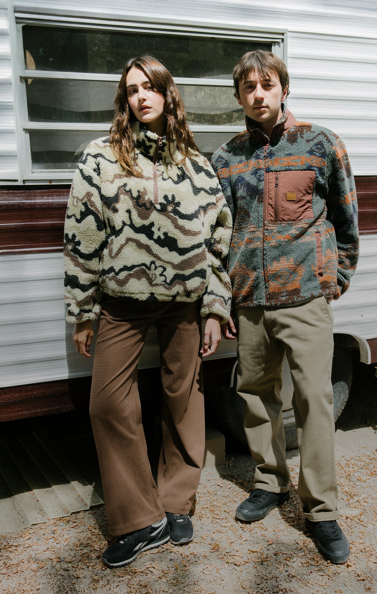 Two people standing in front of a trailer wearing patterned sweaters and pants.