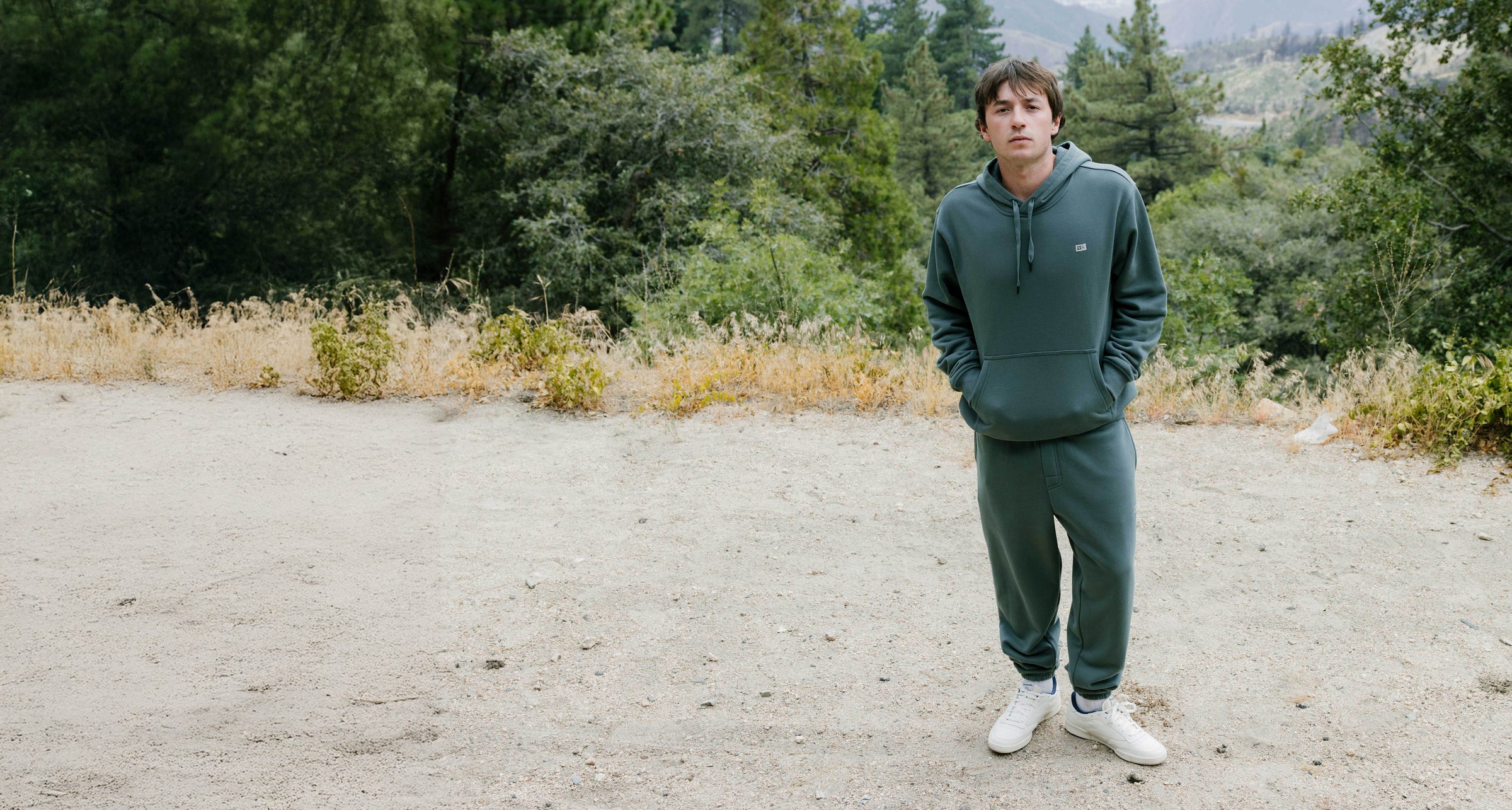 Person wearing a green hoodie and pants standing in a natural setting with trees and open space.