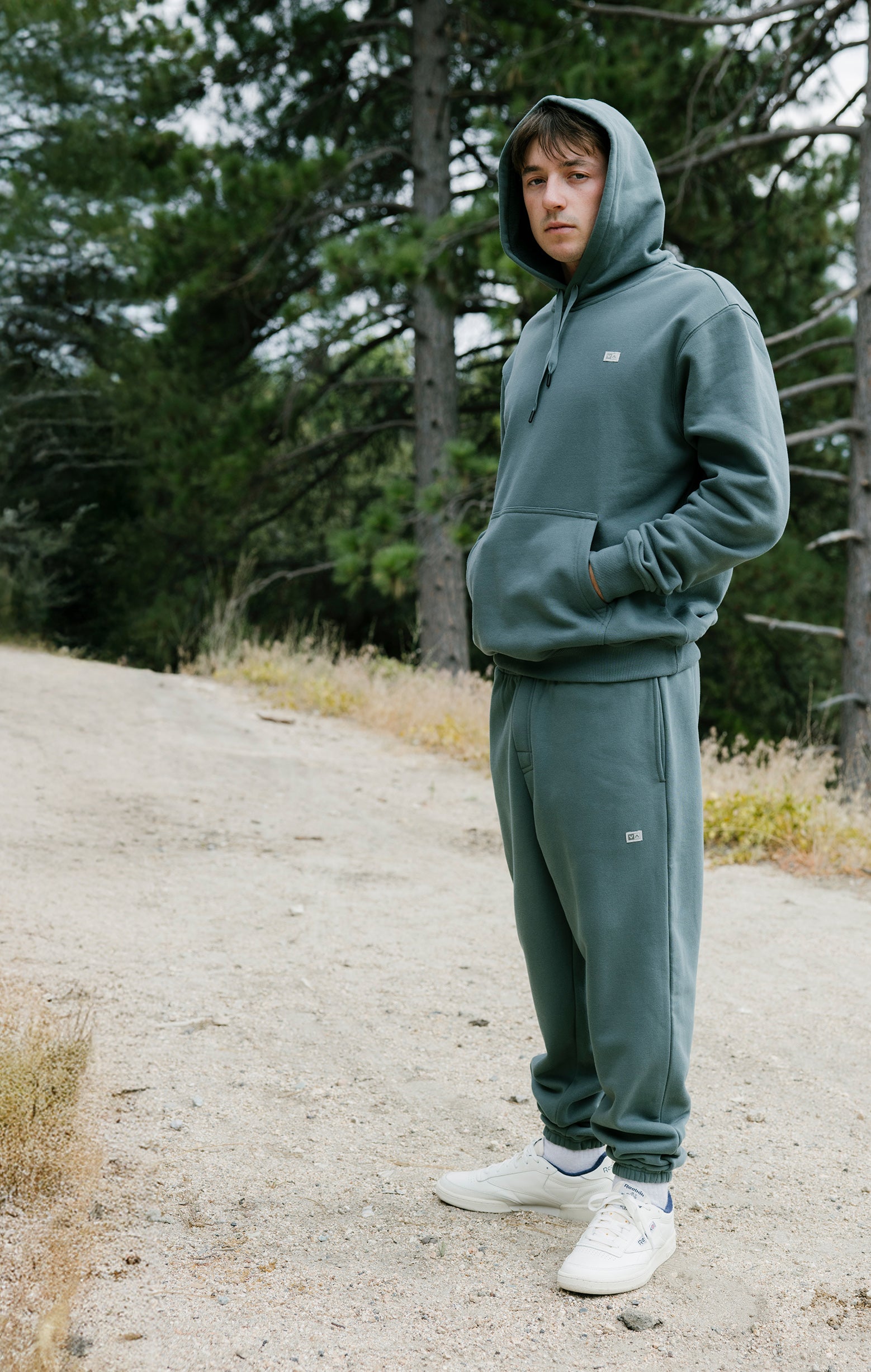 Person wearing a green hoodie and pants standing on a dirt path with trees in the background