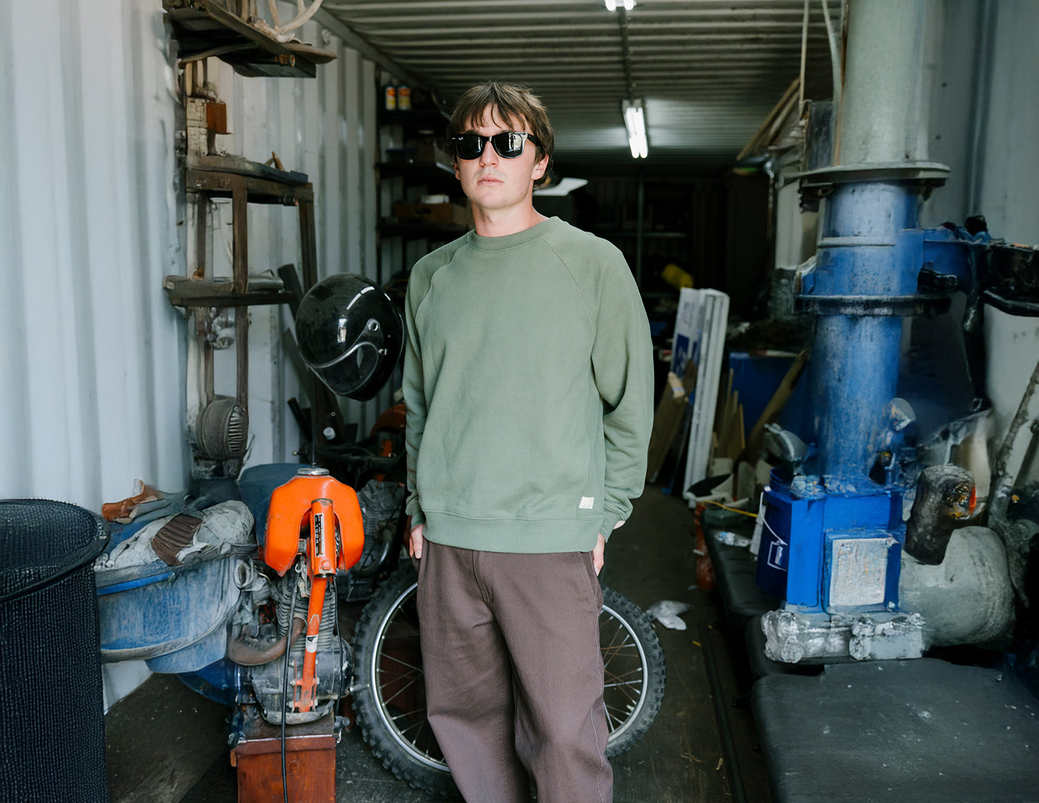 Person wearing sunglasses and a green sweater standing in a workshop with various equipment.