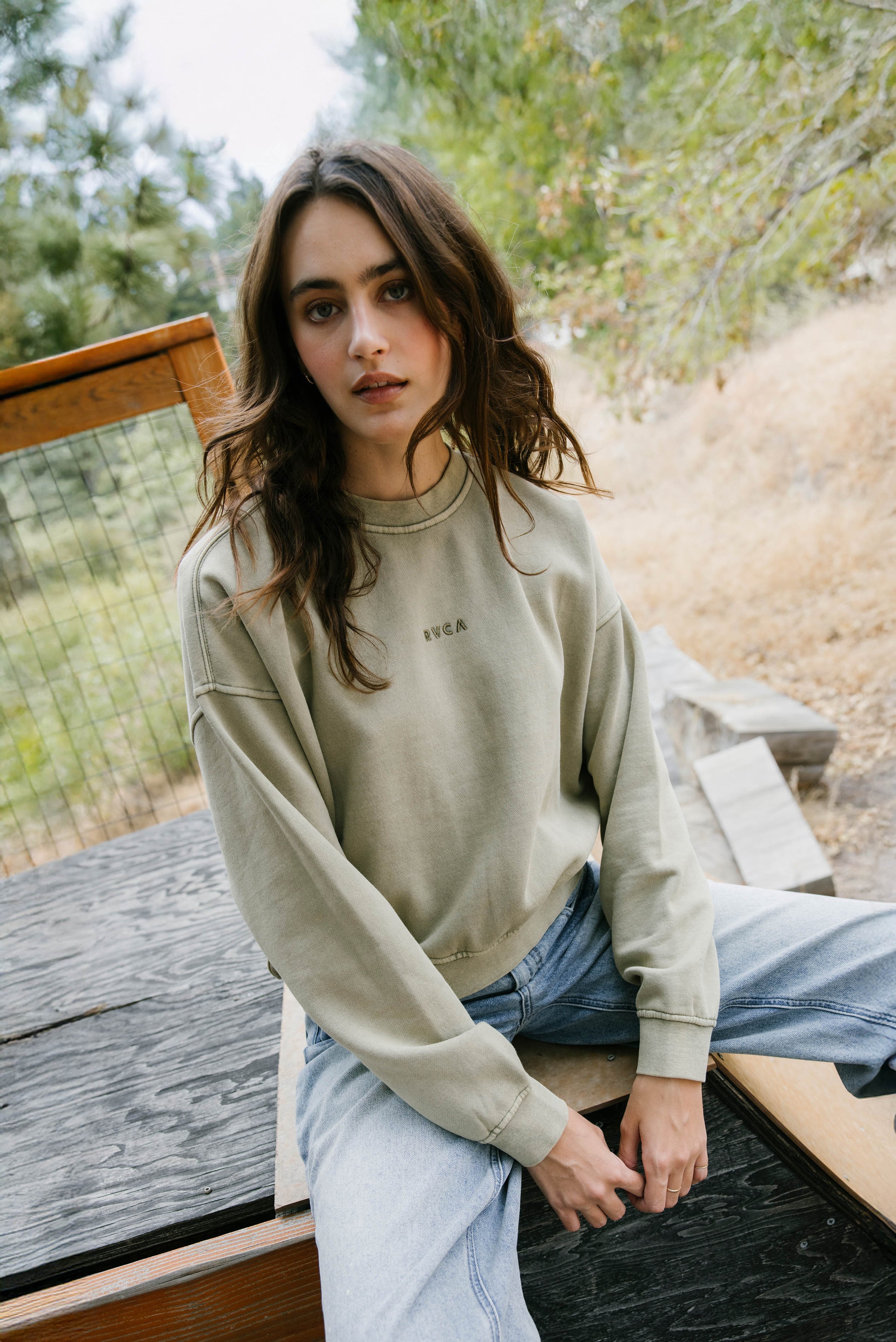 Woman sitting outdoors wearing a beige sweatshirt with text and light blue jeans.