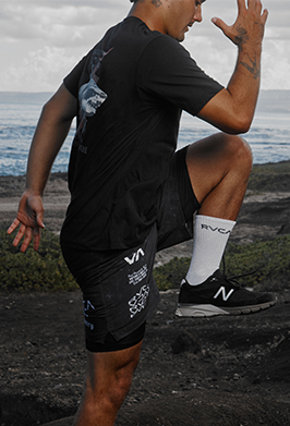 Person wearing black athletic shorts with brand logos, standing on a rocky beach.