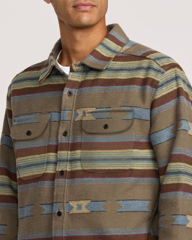 A man wearing the Dayshift Blanket Flannel Long Sleeve Shirt, featuring a multicolored striped and geometric pattern in brown, blue, green, and tan with two chest pockets with flaps and buttons.