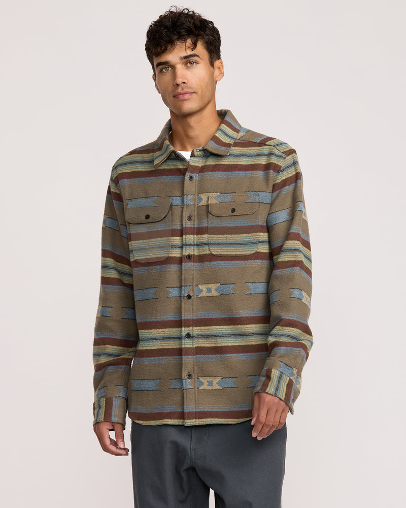 A young man with short curly hair and a neutral expression wears the Dayshift Blanket Flannel Long Sleeve Shirt, featuring brown fabric with horizontal blue, green, and yellow stripes plus geometric patterns, paired with dark gray pants against a plain background.