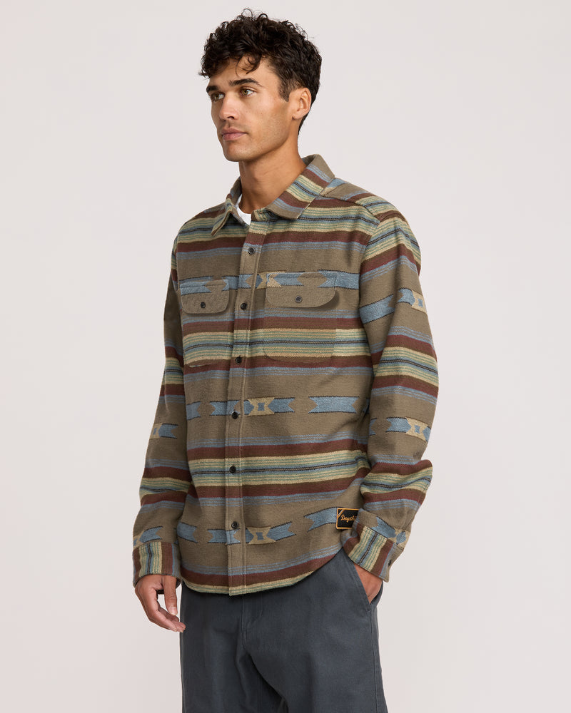 A man wearing the Dayshift Blanket Flannel Long Sleeve Shirt with earth-toned horizontal stripes and geometric designs stands against a plain light background, looking slightly to his right.