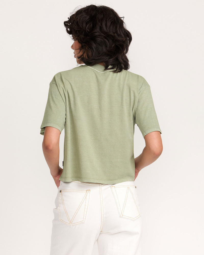 A person with wavy dark hair is shown from the back, wearing the light green Kinney Tee Pocket PTC and high-waisted white jeans with visible seams, standing against a plain light background.