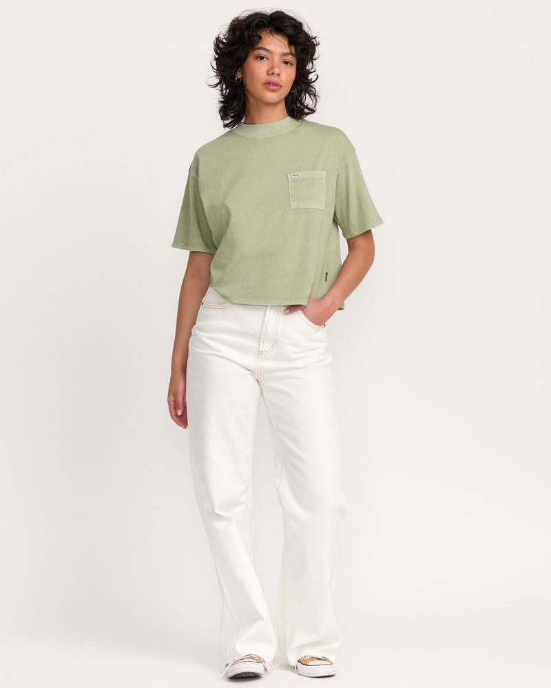 A person with curly hair wears the Kinney Tee Pocket PTC, a loose, light green short-sleeve top with a chest pocket, paired with white wide-leg pants against a plain white background.