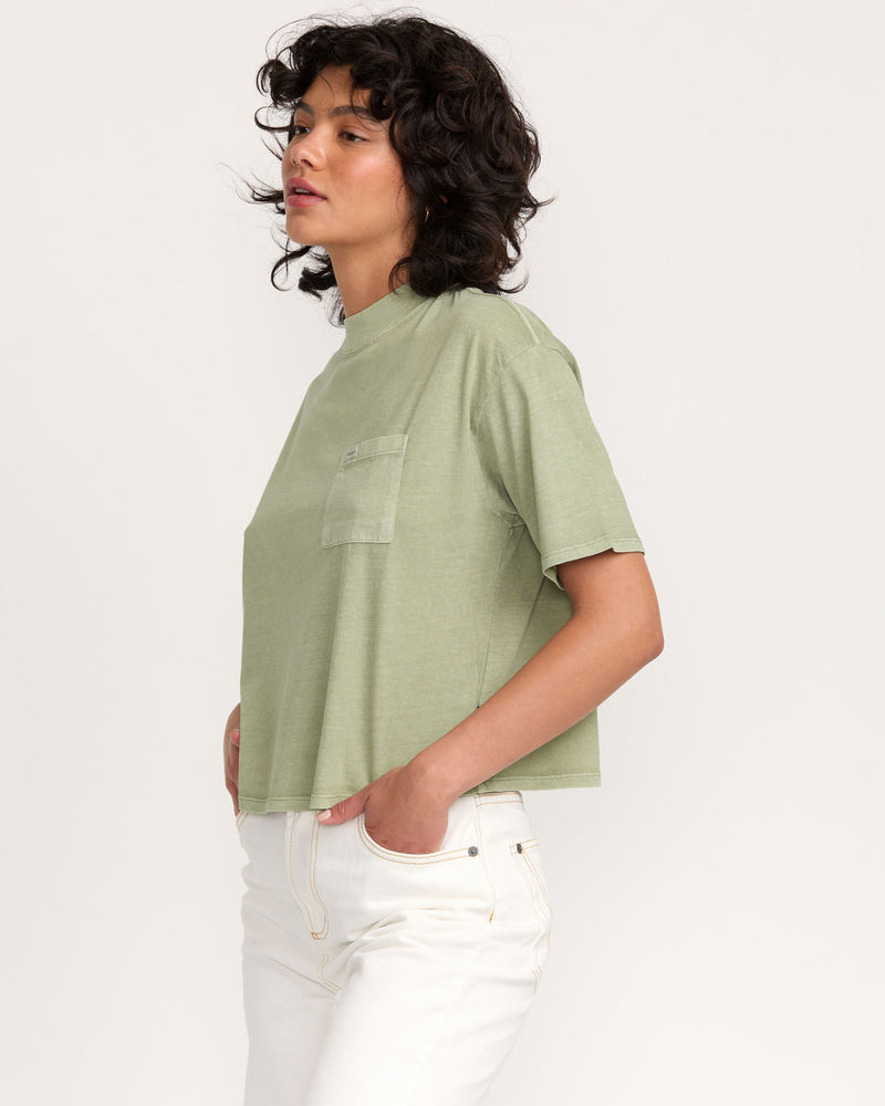 Wearing the Kinney Tee Pocket PTC, a person with curly dark hair and high-waisted white pants stands in profile with hands in pockets against a plain light background.