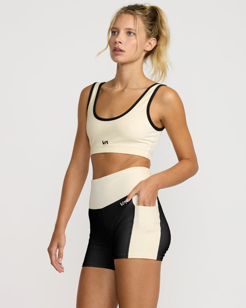A woman wears the VA Essential Pocket Short II, a beige and black high-waisted short, paired with a coordinating beige sports bra. She stands with one hand in her pocket, looking to the side against a plain background.