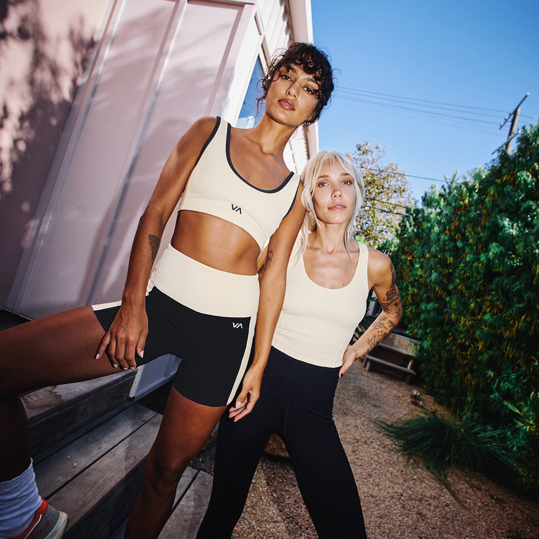 Two women in athletic wear standing outdoors with a building and greenery in the background.
