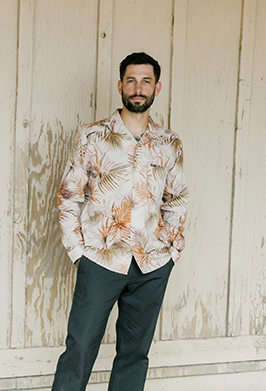Man wearing a floral shirt and dark pants standing against a wooden wall.