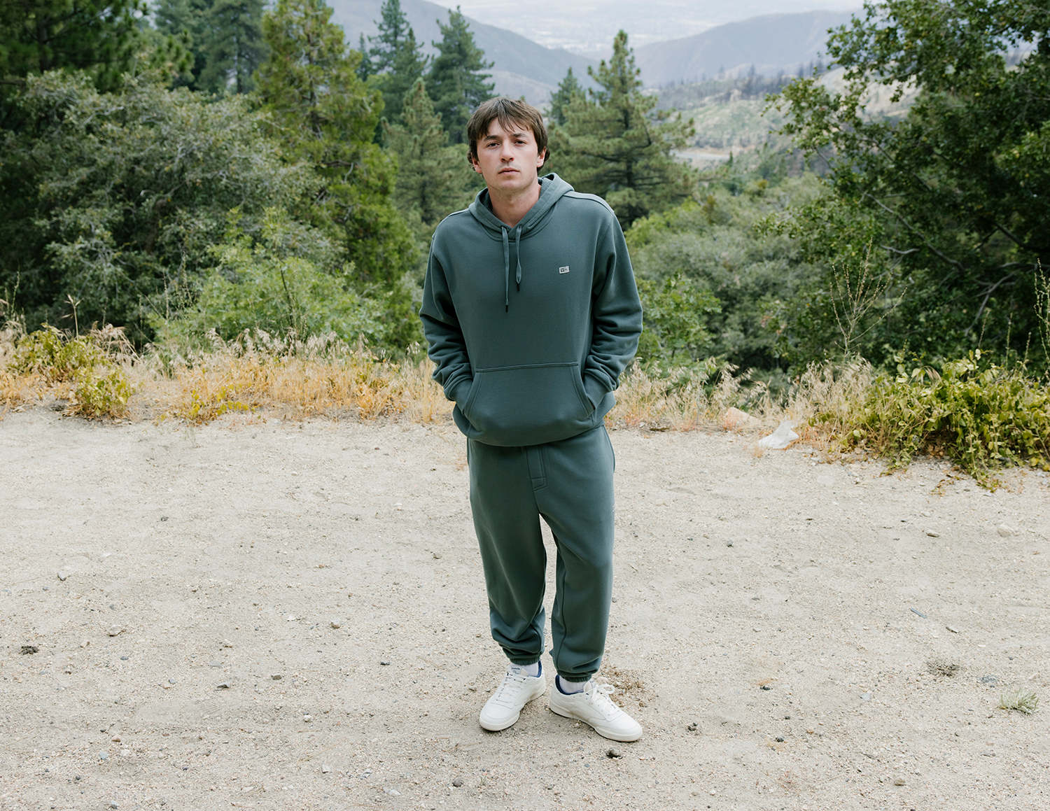 Person wearing a green hoodie and pants standing on a gravel path with trees and mountains in the background