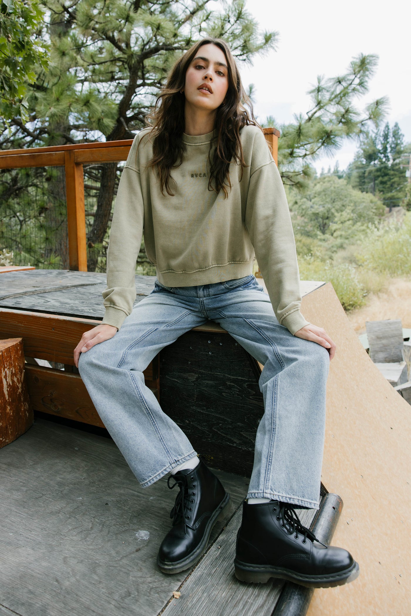 Woman sitting outdoors wearing a beige sweater, blue jeans, and black boots.