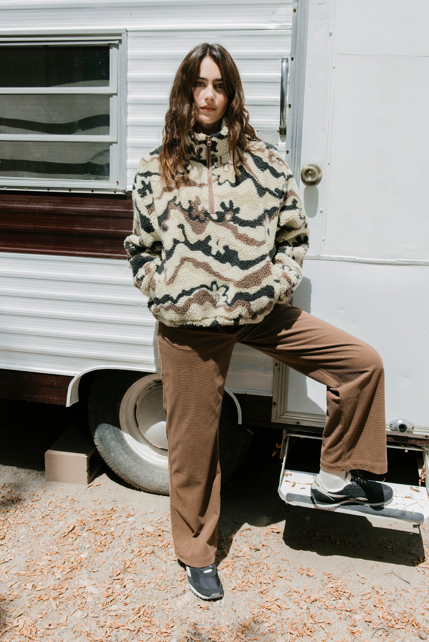 Person wearing a patterned sweater and brown pants standing in front of a trailer.