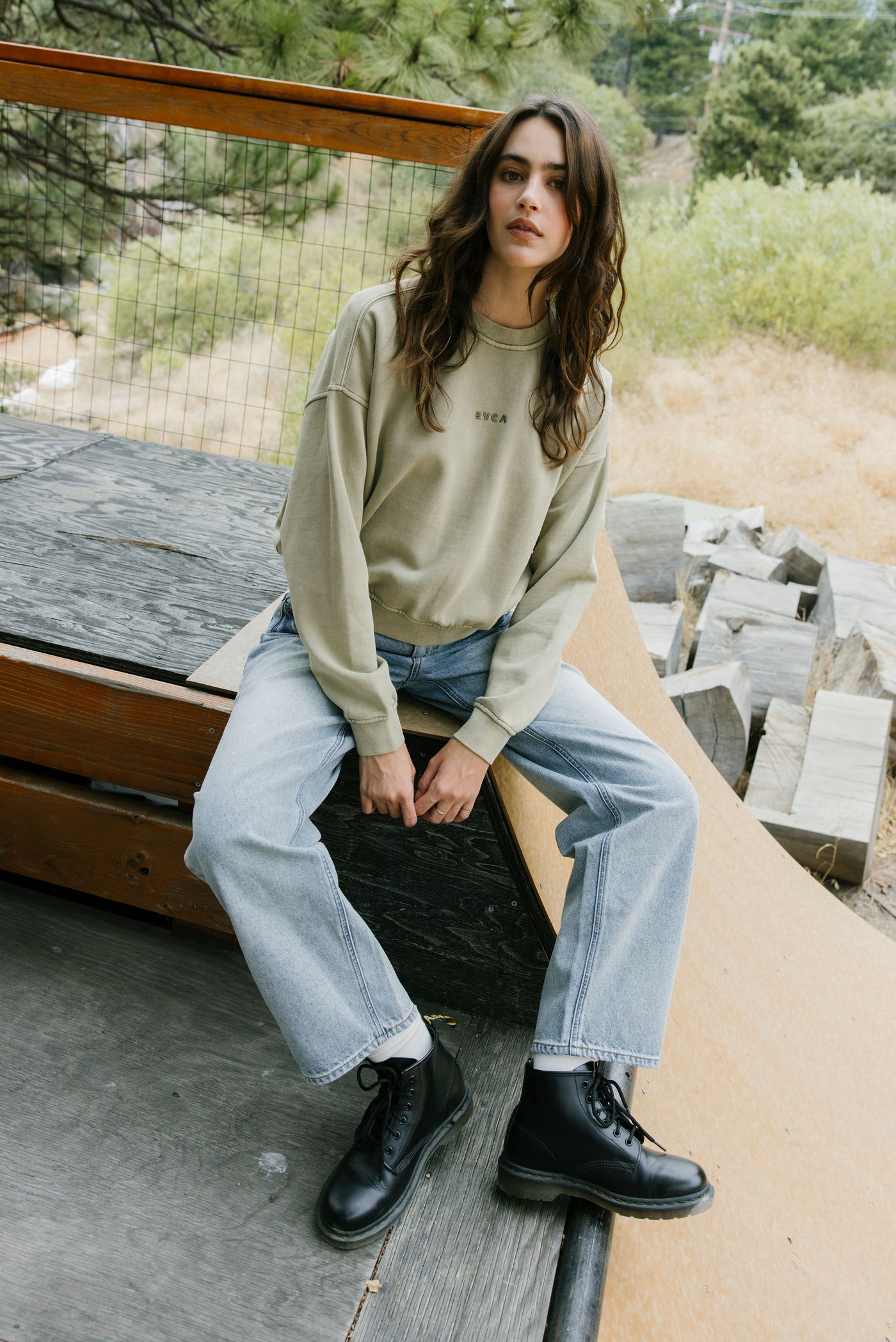 Woman sitting outdoors wearing a beige sweater, light blue jeans, and black shoes.